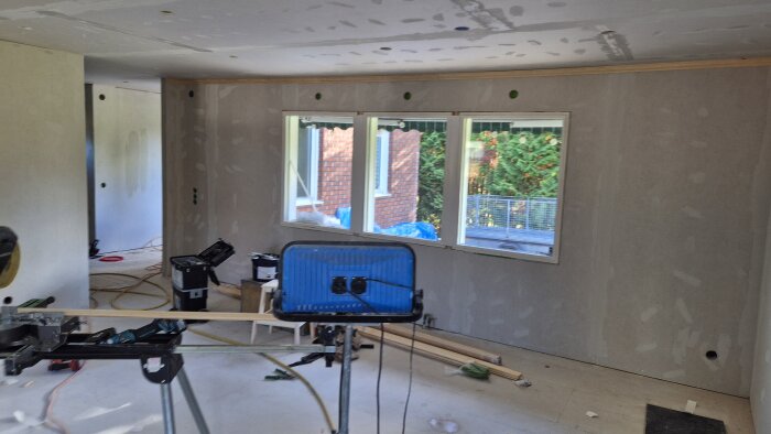 Renoveringsprojekt med gipsade väggar och tak, bygglampor och verktyg. Stora fönster visar utsikt mot trädgård och tegelvägg.