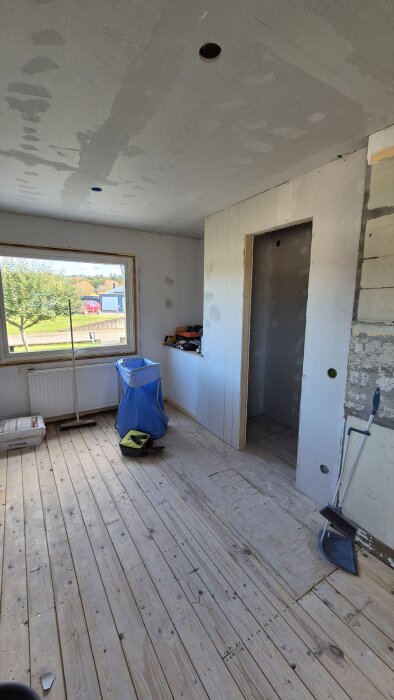 Renoveringsprojekt med spacklade gipsskivor, träplankgolv och fönster. Verktyg och byggmaterial syns i rummet.
