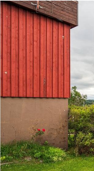 Röd träpanel på husvägg ovanför putsad grund, med grönska nedanför och molnig himmel i bakgrunden.