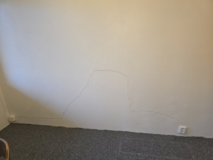 Sprucken vägg i källarrum med synliga sprickor, eluttag och heltäckningsmatta, inför renovering och lagning med ankarmassa och spackel.
