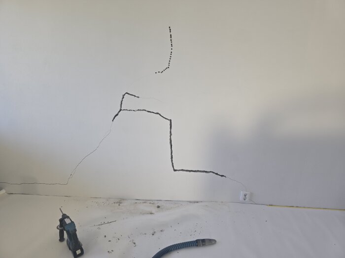 Vägg med stora sprickor och borrverktyg, förberett för reparation med ankarmassa och lagningsbruk. Synlig dammsugarslang och eluttag på golvet.