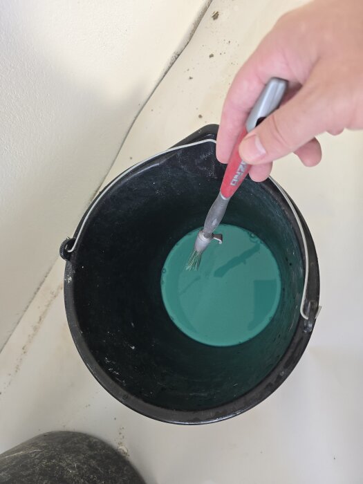 Svart hink med blågrön primer och pensel, använd för förberedelse vid reparation av vägg i källarens sovrum.
