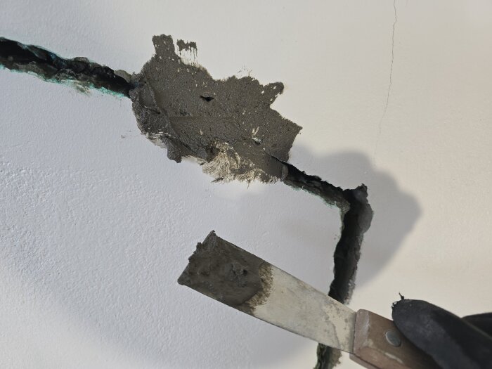Lagning av spricka i vägg med lagningsbruk. En spackelspade används för applicering. Sprickan är bred och tydligt synlig.