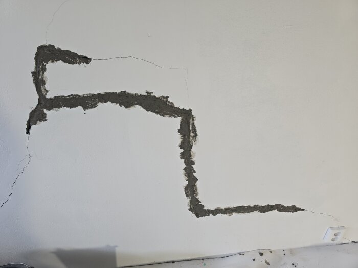Sprickor i betongvägg lagade med ankarmassa, synlig svart massa applicerad längs spricklinjer på vitmålad yta. Fragment av byggmaterial på golvet.