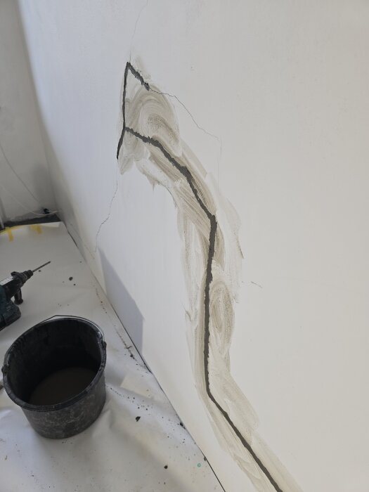 Spricka i vägg med påbörjad lagning med ankarmassa och lagningsbruk, böjda spricklinjer och spann med verktyg på golv.