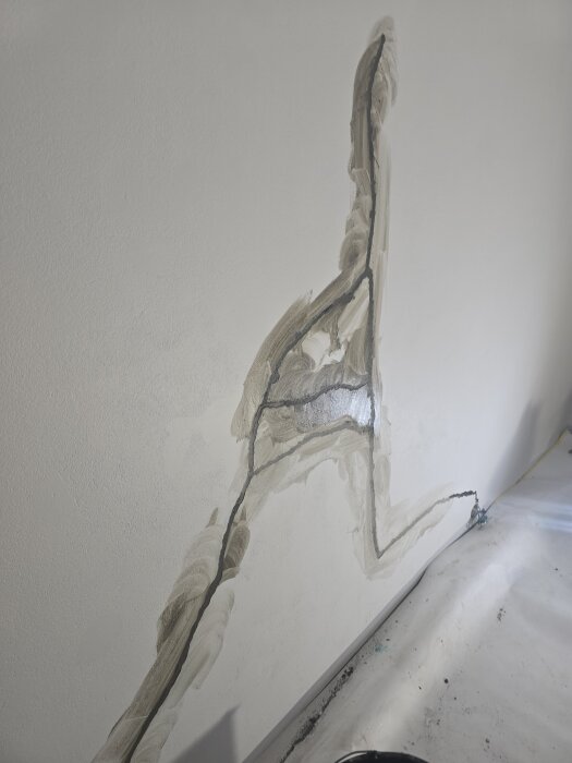 Sprickor i källarvägg lagade med ankarmassa. Ytan förbereds för spackling och målning, med synligt reparationsarbete i grått bruk. Plasten täcker golvet.