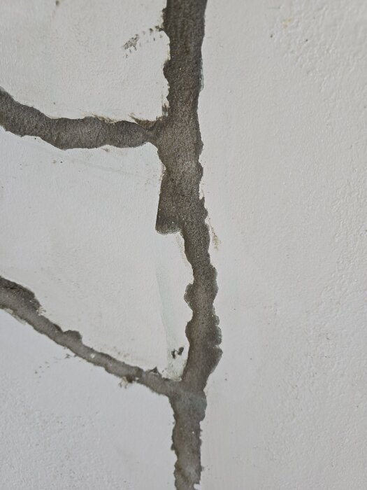 Bild på en vitmålad vägg med stora sprickor som täcks med lagningsbruk, del av ett renoveringsprojekt för att förstärka väggens struktur.
