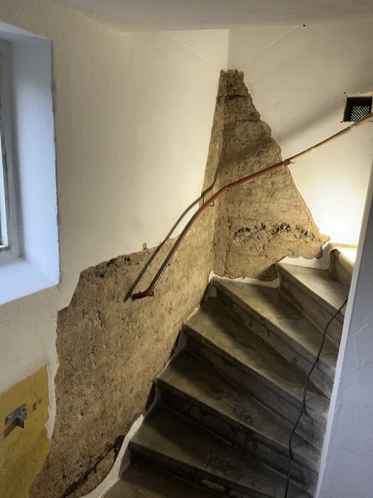 Trappa med delvis bortbilad gammal lagning på vägg, med synlig brun puts och träräcke; arbete förberett för omputsning med kalkbruk.