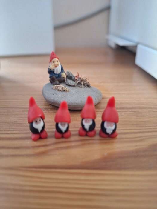 Små tomtefigurer med röda luvor, en större sitter på en sten omgiven av svampar och kottar, fyra mindre tomtar står i förgrunden på ett träbord.