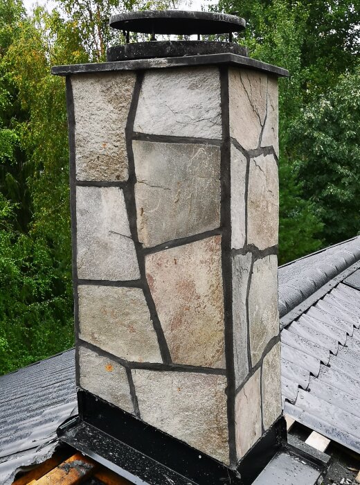 Skorsten klädd med Glavaskiffer, belägen på ett tak med omgivande grönska i bakgrunden.