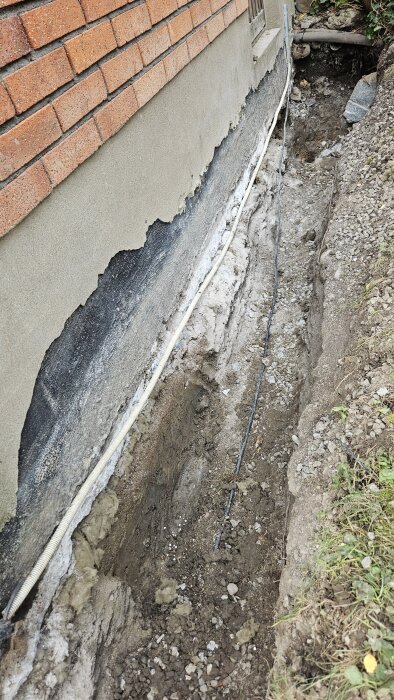 Grävd ränna längs en husvägg med synlig stenull och rör, förberett för isolering av bottenplatta.