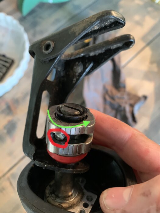 En närbild på en hand som håller en teknisk komponent med en justeringsmekanism, markerad med en röd cirkel och grön pil, på ett träbord.