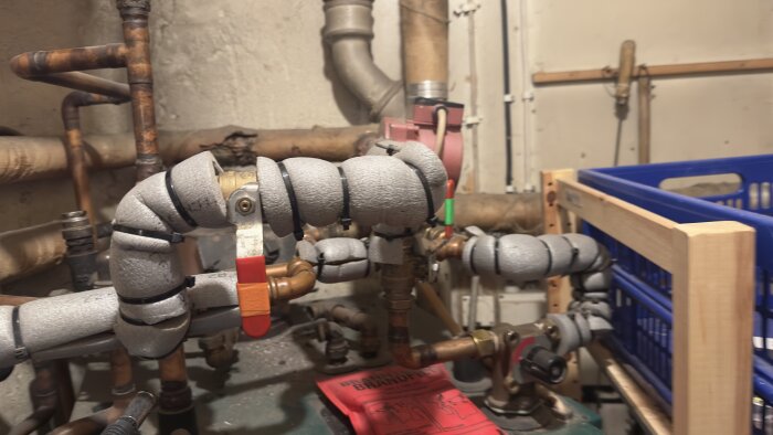 Vattenburet värmesystem i pannrum med kopparrör och isolerade kulventiler i fokus. En blå korg och brandfilt syns i bakgrunden.