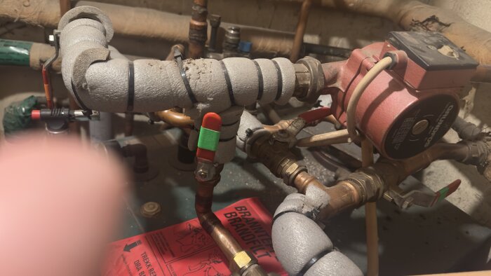 Kulventiler och rörledningar i ett pannrum med värmesystem, synlig pump märkt "Grundfos" och isolerade rör. En röd brandsläckare syns delvis.
