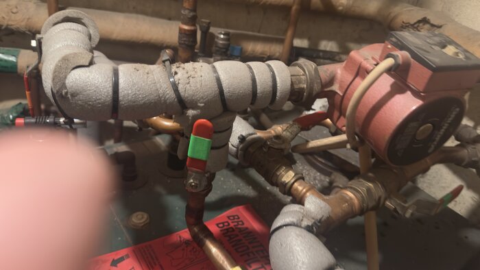 Kulventiler och rör i ett pannrum med isolering och en cirkulationspump syns. En brandsläckningsinstruktion ligger nedanför.