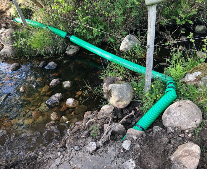 Gröna dräneringsrör ligger över mark och sten bredvid ett grunt vattendrag, omgivna av grönska och ett enkelt stängsel.