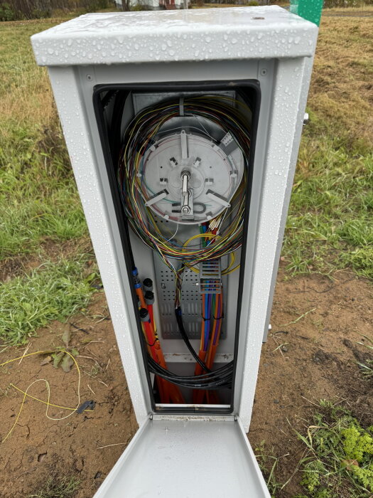 Öppen fiberoptikbox utomhus med synliga kablar och fukt på ytan, placerad på gräsig mark.