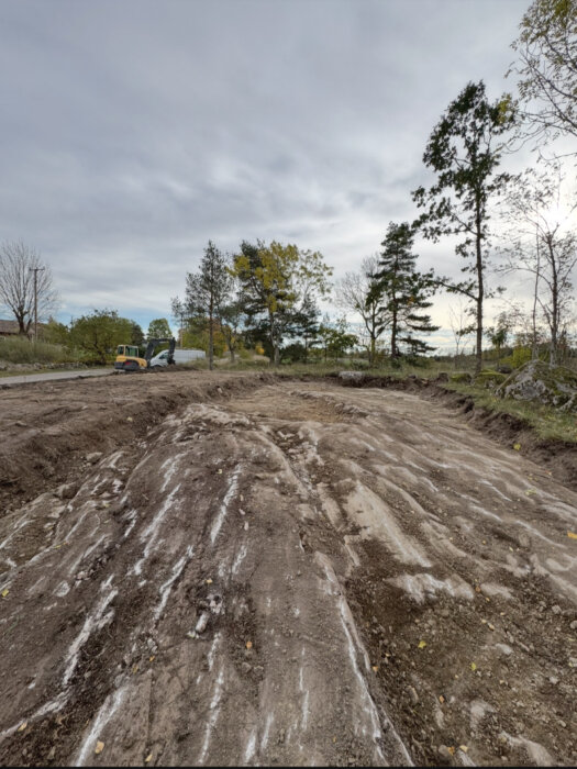 Markarbete på tomt med synligt berg under, omgivet av några träd i bakgrunden och en grävmaskin synlig till vänster.