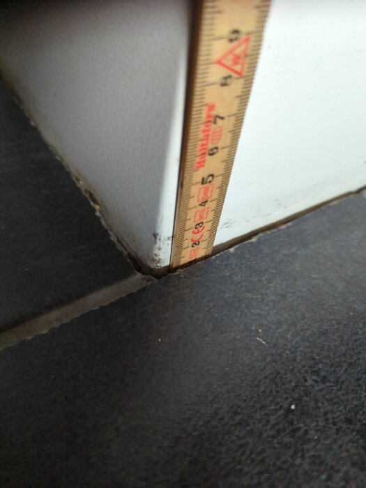 Tumstock mäter avståndet mellan en vit vägg och ett svart klinkergolv, potentiellt problem vid avmontering av fläkt pga kakel.