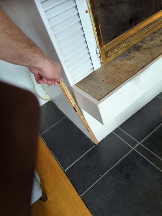 Hand håller tumstock vid vit kåpa intill svart kakelgolv för renovering.