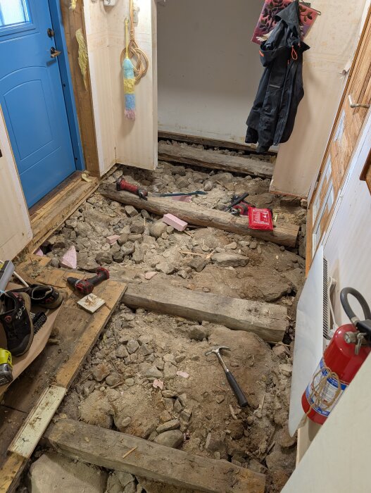 Rummet visar ett golv under renovering med exponerade bjälkar, verktyg på marken, en dörr i bakgrunden och upphängda kläder.