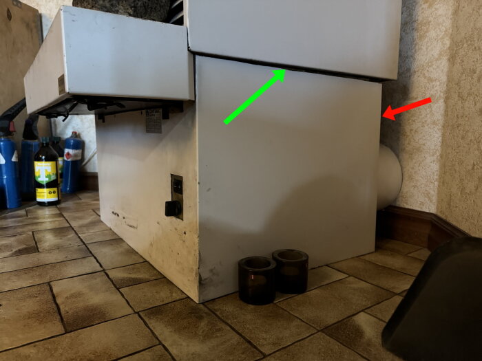 Plåtkåpa vid kamin, fastsatt i klinkergolv. Grön pil visar slipad överdel; röd pil indikerar åtdragna skruvar. Synliga ytfläckar på plåt och golv.