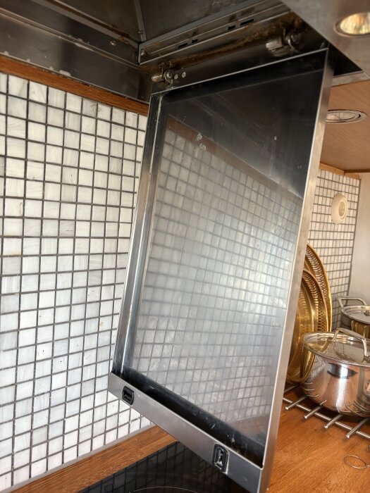 Öppen köksfläktslucka monterad med skruvar, svåråtkomliga för att avlägsna. Bakgrund med kaklad vägg och köksbänk med kastruller.