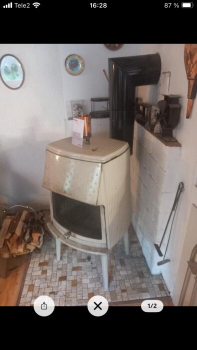 Gammaldags plåtkamin med glaslucka, ljus färg, står mot vit vägg, ansluten till svart rör, omgiven av ved och verktyg.