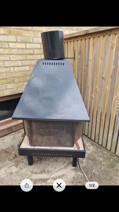 Stor plåtkamin med rektangulär bas och lutande topp på uteplats; placerad framför en tegelvägg och trästaket.