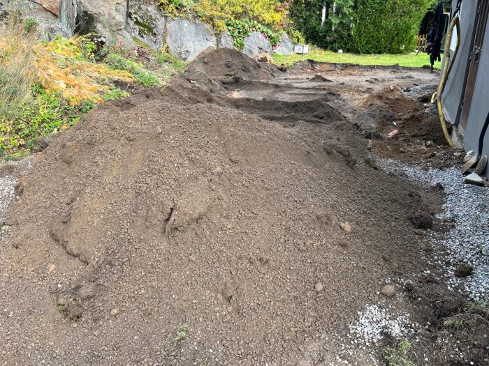 Hög av jord och kross på en grävplats vid sidan av ett hus, omgiven av grönska och stenar.
