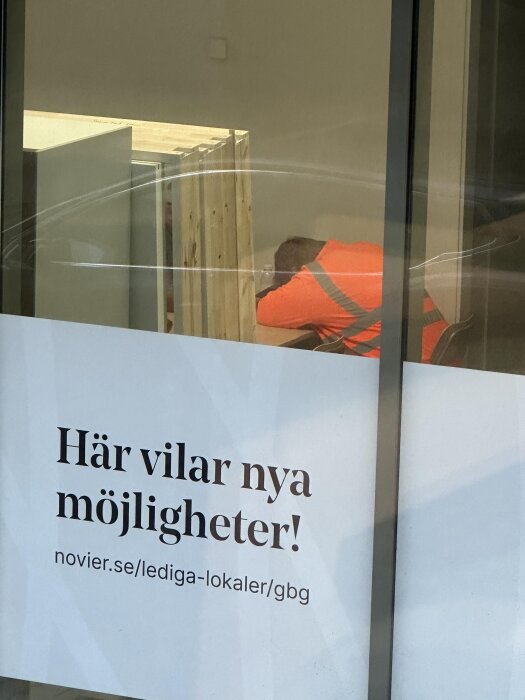 Byggarbetare i orange klädsel vilar vid ett bord inomhus, sett genom ett fönster med texten "Här vilar nya möjligheter!".