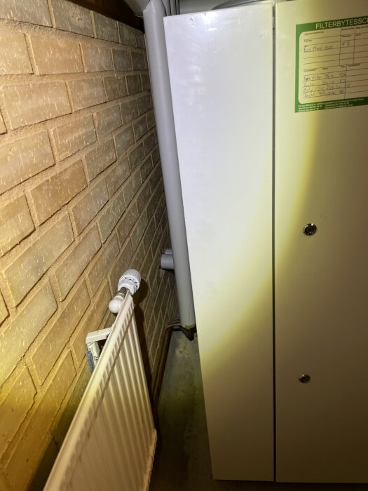 Teknikrum med tegelvägg, stort fläktaggregat och rör bakom radiator. Begränsat utrymme för rördragning och arbete.