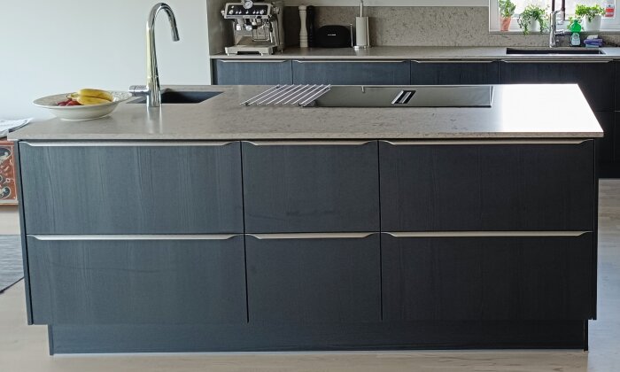 Köksö med mörka skåp, inbyggd häll med fläkt, diskho och kran. Kaffemaskin och skål med frukt syns i bakgrunden.