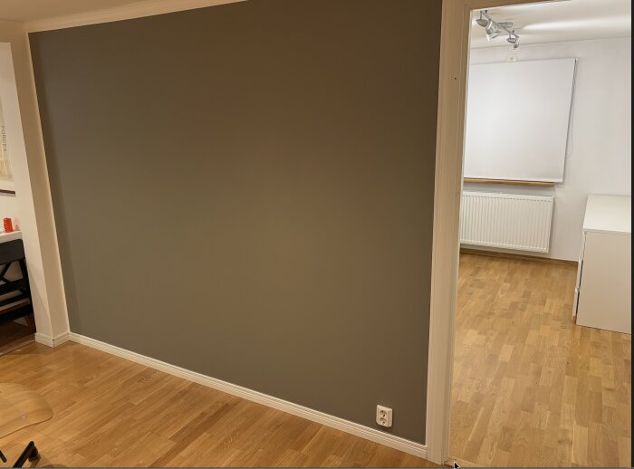 Inomhusvägg i ett rum, med trägolv och en vit dörrkarm, visar en slät, grå yta. Det finns en eluttag vid golvet.