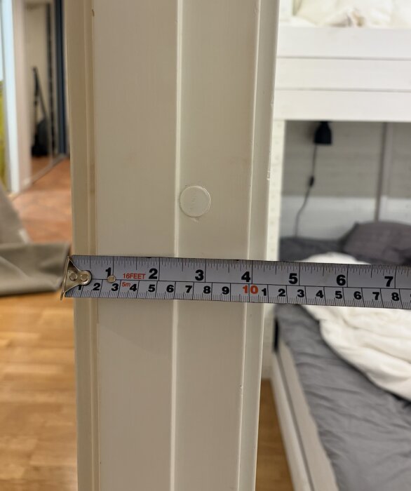 Mätband håller på att mäta tjockleken på en innervägg i ett rum, med en säng i bakgrunden.