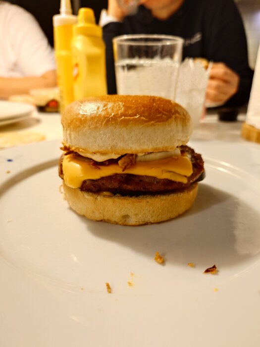 Hamburgare med ost, dubbel lök, ketchup, senap, saltgurka och sourcream pepper på ett vitt fat, med senapsflaskor i bakgrunden.