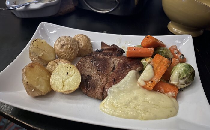 Fläskkarré med ugnsstekt potatis, brysselkål och morötter serveras på en vit tallrik tillsammans med sås.