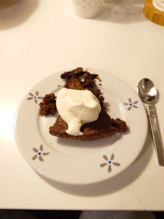 En bit kladdkaka med grädde på en blommönstrad tallrik, placerad på ett vitt bord bredvid en sked.