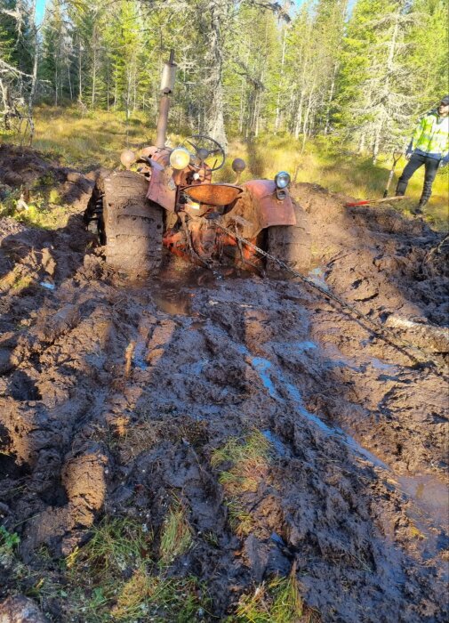 En gammal traktor fast i lerig mark i skogen under dagsljus, omgiven av träd och en person i bakgrunden.