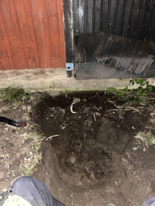 Grop grävd vid en svart och röd vägg, invid en dörr med skylten "NB" synlig.