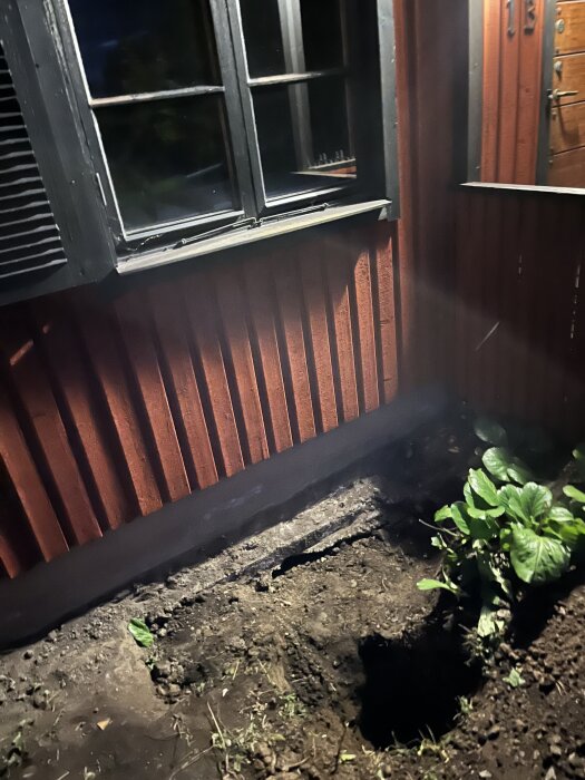 En grävd grop framför ett rött trähus med svart fönsterkarm och en grön buske bredvid.