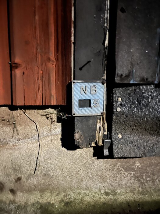 Skylt med texten "NB" ovanför en siffra "5" på en vägg bredvid ett rött träpanel, bredvid grävt jordområde.