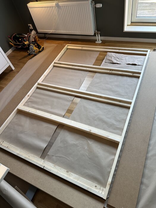 En träram ligger på golvet redo att monteras som en del av ett sängskåp, omgiven av verktyg och material i ett rum.