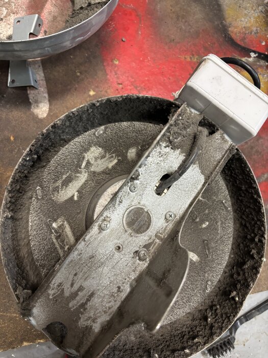 Sliten betongblandare med smuts och damm på insidan, omgiven av en röd och brun yta i en verkstadsmiljö.