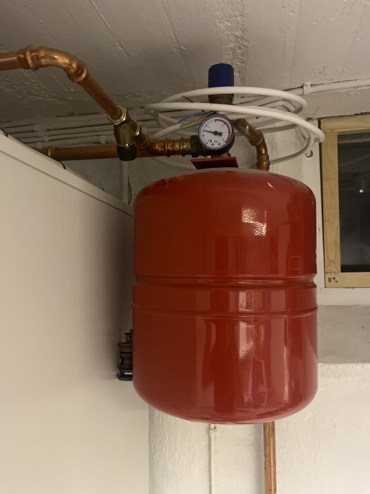 Tryckmätare och röd trycktank med kopparrör och svart ventil i ett värmesystem.