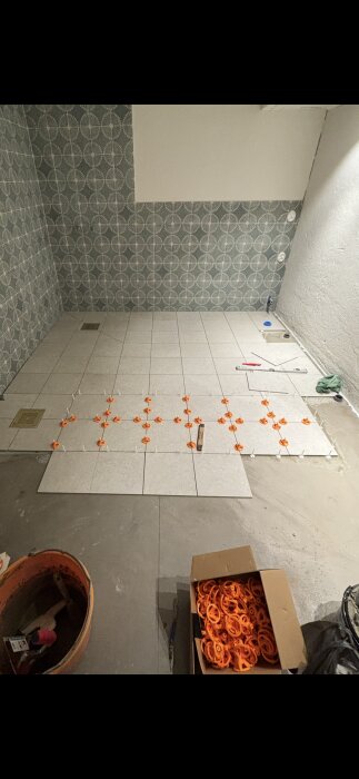 Kakelgolv i färd med att läggas, med orange kakeldistanser. Delvist klar vägg med dekormönstrat kakel. Verktyg och material syns.