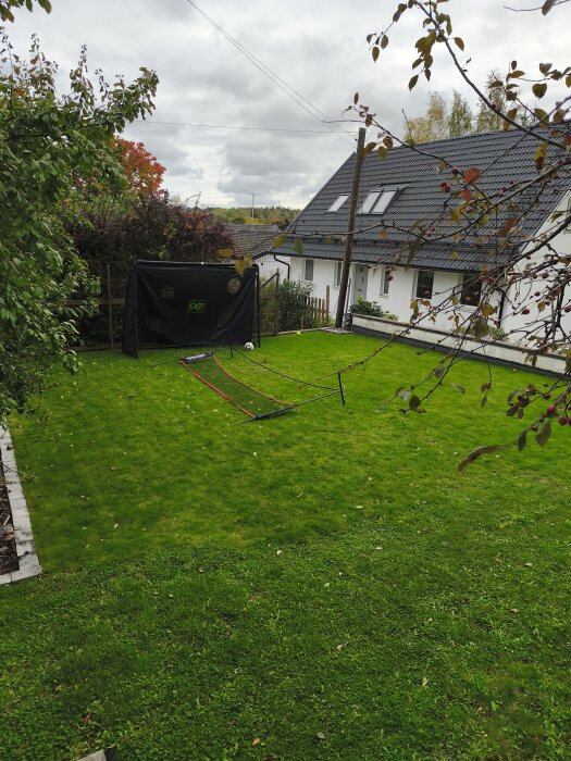 Gräsmatta med badminton- och fotbollsmål, omgiven av träd och buskar, med ett hus och en trästolpe i bakgrunden.