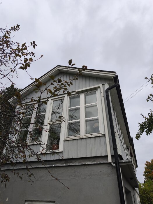 Fasaden av ett hus med vita fönster och grå träpanel, omgiven av trädgrenar, under en molnig himmel. Eventuell plats för utebelysning.