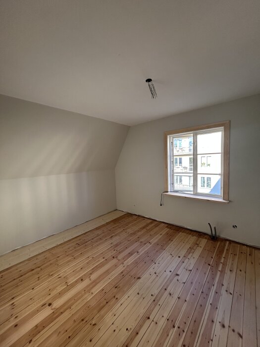 Nyrenoverat rum med trägolv i olika nyanser. Ett fönster syns på höger sida, och taket har en oskarv sladd hängande.