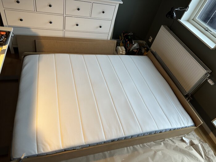 Murphy Bed i nedfällt läge utan golvsockel och knoppar, visar en vit madrass i ett träram, omgiven av verktyg och en vit byrå.